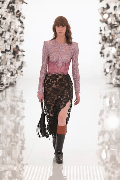 Powder-pink lace top with padded shoulders and a scalloped waist. Black floral lace skirt over nude lining, slit cut high at one thigh. Black fringed pouch on a chain strap. Black knee boots with a rust cuff and square buckles. Crossing a hotel atrium after midnight with the bouquet wrapper still in hand.