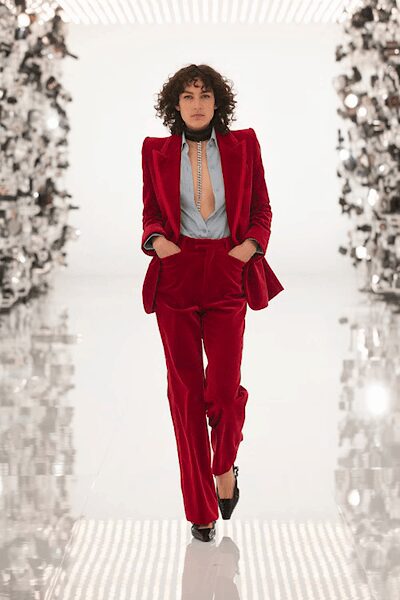 Crimson velvet blazer with peaked shoulders and satin lapels. Pale blue blouse plunged open, held by a narrow chain placket. Black choker tight at the throat. Matching velvet trousers breaking softly over the shoe. Black pointed heels. Walking into a hotel bar before anyone else from the wedding arrives.