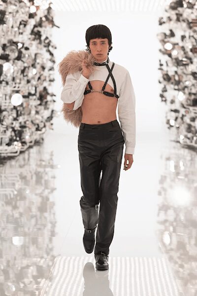 White cropped shirt hanging open, one cuff loose at the wrist. Black leather harness buckled over the bare chest and neck. Blush shaggy stole slung over one shoulder. Black leather trousers with a lean straight leg. Black loafers with metal bit hardware. Walking into a mirrored corridor where the heating has failed.