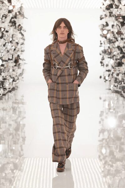 Narrow oxblood neck scarf knotted close, tails lost under the lapel. Double-breasted brown plaid blazer cinched with a silver horsebit harness. Matching plaid trousers breaking softly over the shoe. Dark leather loafers with a dull metal bit. Walking through a hotel arcade before the breakfast trays are cleared.