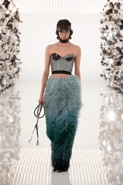 Black quilted cap with ear-covering flaps and a snug chin strap. Silver beaded corset top, black cups and a narrow grosgrain waist. Column skirt shagged in feathers, fading from pale blue to deep teal. Beaded loop handle with loose black ribbon tails. Crossing a winter garden tunnel after midnight champagne.