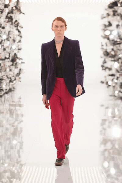 Deep aubergine velvet blazer with peaked lapels and a long clean line. Black draped V-neck top disappearing into the jacket opening. Hot pink fluid trousers, pressed crease running straight to the shoe. Short crystal collar necklace. Dark square-toe shoes. Walking into a hotel ballroom before the wedding guests arrive.