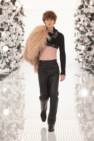 Blush shaggy fur stole slung off one shoulder. Black one-sleeve crop top with ring hardware at the throat. Black leather trousers, creased through the front and slightly flared. Square-toe black boots. Whatever you wear home when dawn catches you under supermarket fluorescents.