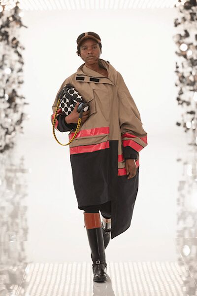 Brown cap with a pale center stripe down the crown. Tan utilitarian poncho, cut with fluorescent reflective bands and black tabs. Single black glove. Yellow rope strap looped from the hand. Black knee boots, one leg capped with a chestnut shin guard. Crossing a flood barrier after the sirens stopped.