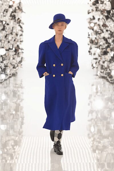 Cobalt felt hat with a flat brim and shallow crown. Cobalt double-breasted coat, broad lapels and gilt buttons in pairs. Black and grey argyle knee socks. Black leather lace-ups with a rounded toe. Crossing a hotel atrium before breakfast, suitcase wheels already clicking behind you.