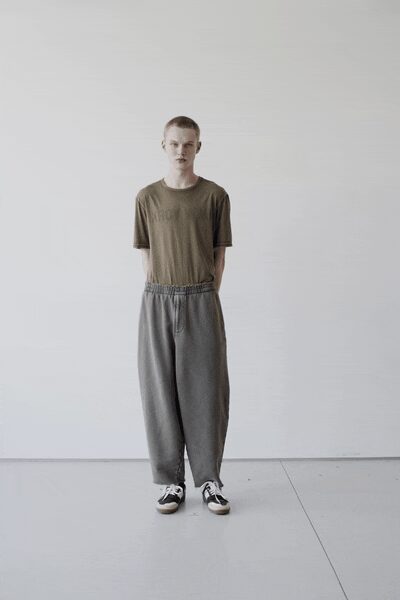 Faded olive jersey tee, the chest lettering nearly rubbed away. Ash grey drawstring trousers cut very wide, waistband gathered in soft puckers. Black-and-white low-top sneakers with thick off-white rubber toes. First hour after moving day, standing in an apartment with no furniture yet.