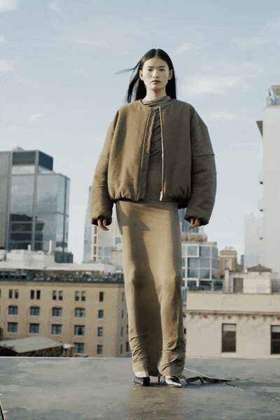 Cropped taupe bomber, heavily padded with a twisted trim running off-center. High-neck underlayer in close beige knit. Column skirt in putty jersey, pooling and wrinkled around the ankles. Black-and-white pointed mules. Loose strap dragging beside one foot. Wind on the roof after office lights have started going out.