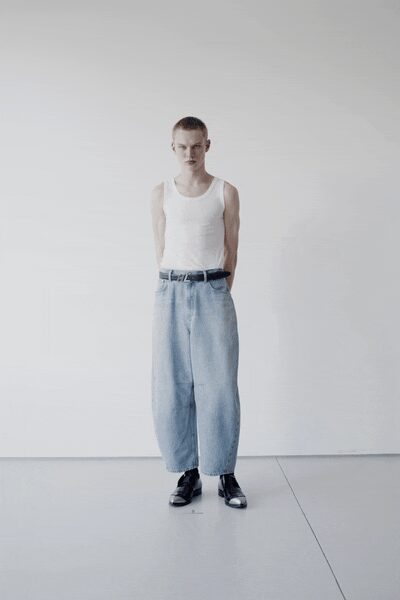 White ribbed tank, cut close with deep armholes. Black leather belt with a plain silver buckle. Bleached wide-leg jeans, ballooning out from sharp front pleats. Black leather shoes with square toes and thick soles. Sunday laundry room, someone else's detergent still in the air.