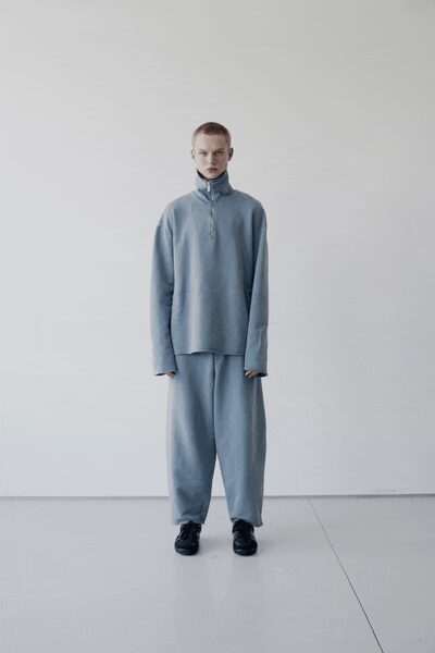 Powder-blue knit top with a funnel neck and half-placket seams. Matching wide knit trousers falling loose and straight from the hip. Black lace-up shoes with a blunt, rounded toe. Morning in a cold studio before the radiators have caught up.
