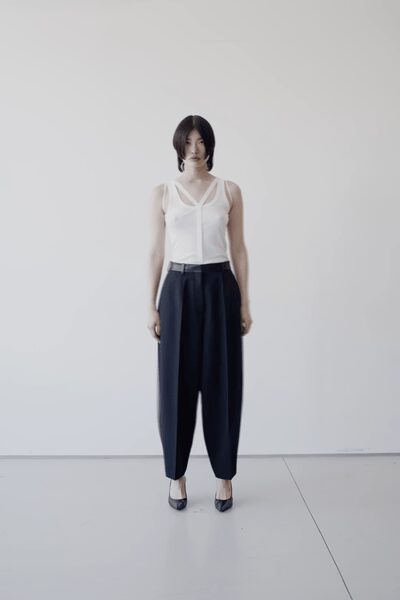 Ivory knit tank with a sheer yoke and narrow V-trim framing the chest. Inky high-waisted trousers cut full through the leg, tapering softly at the ankle. Black patent heels with a rounded throat. Early appointment in a bare studio before the samples are unpacked.