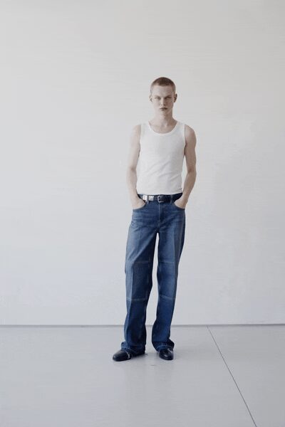 White ribbed tank, cut close with scooped armholes. Black leather belt with a bright rectangular buckle. Dark indigo jeans, whiskered at the hips and breaking over the shoes. Black square-toe shoes with a blunt, glossy front. Empty apartment afternoon, still deciding what stays in the boxes.