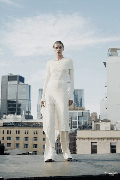Ivory jersey top wrapped diagonally across the shoulders, one cape-like panel falling back. Fluid ivory trousers with a long split overlay drifting from one hip. Pointed white boots peeking out under the hem. Wind on a rooftop after the office towers have emptied.