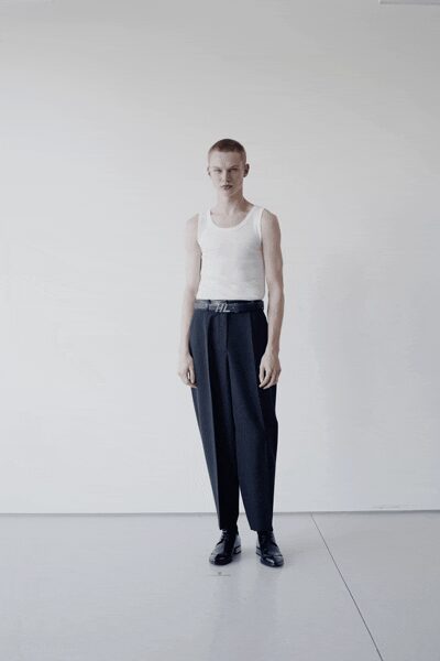 White ribbed tank, cut close with a slightly stretched neck. Dark pleated trousers ballooning through the leg, tapering hard at the ankle. Black belt with a brushed metal initial buckle. Black leather lace-ups with a square, light-catching toe. Morning in an empty rental studio before the steam heat kicks in.