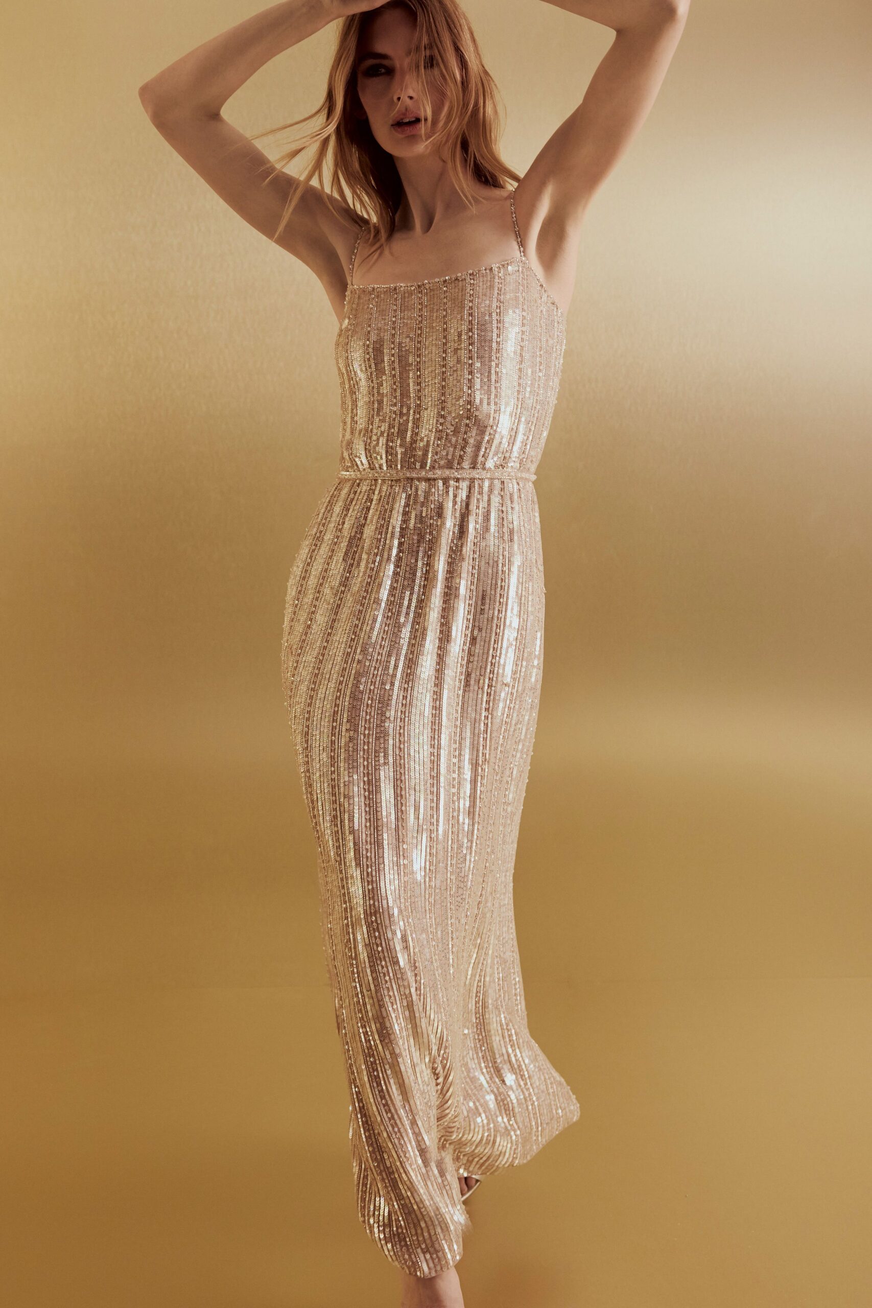 Blush column gown densely striped with sequins, spaghetti straps and a cord-thin waist seam. Pale pointed heels peeking from the swaying hem. Hotel ballroom at noon, before anyone has turned down the chandeliers.