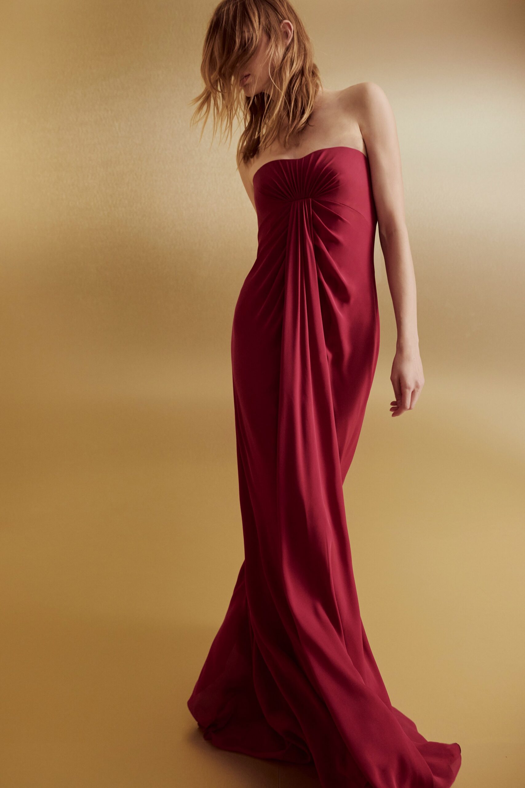 Strapless claret column gown in fluid satin, sunburst ruching knotted off-centre at the bust. The hotel corridor after midnight, heels in one hand, keycard missing again.
