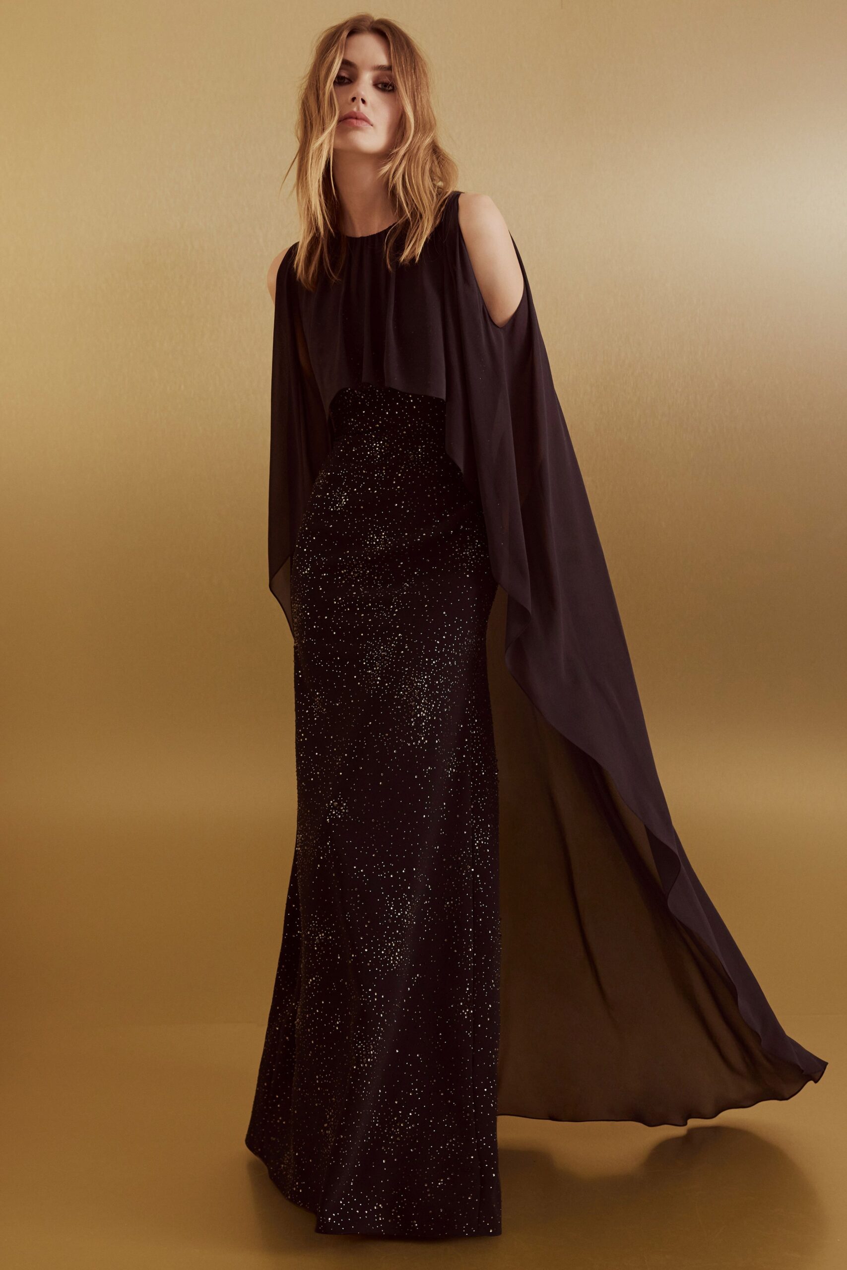 Black chiffon cape gown with cold-shoulder cutouts and a gathered jewel neck. Floor-length black column skirt scattered with fine glitter like dusted ash. Crossing a hotel ballroom after midnight, hem catching the last light.