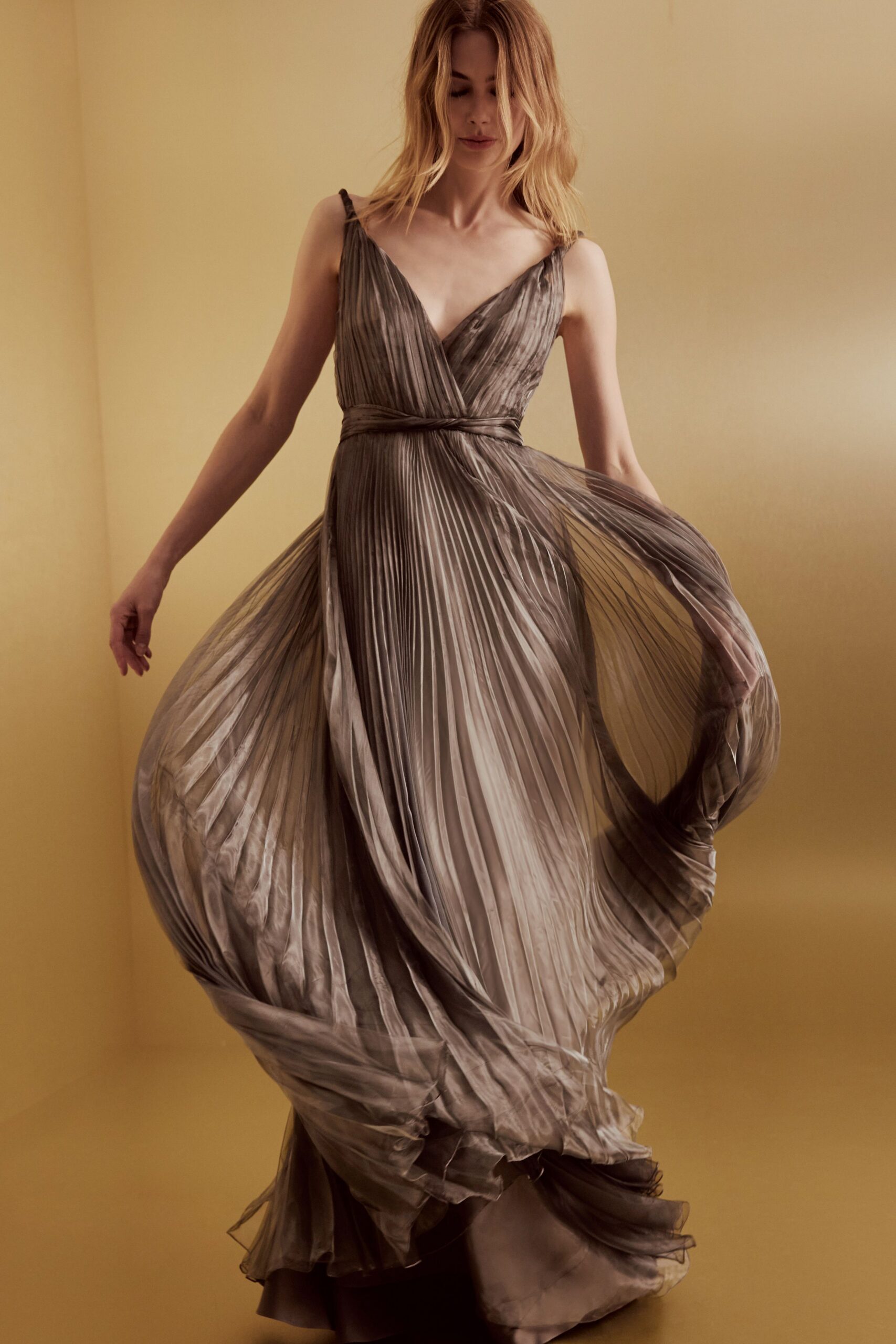 Taupe satin-gleam gown, finely pleated from narrow straps to the floor. Twisted self-fabric band cinching the wrap bodice at a low empire line. Crossing a hotel ballroom after the speeches, skirt still holding the turn.