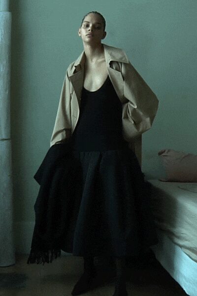 Sand trench coat shrugged open, sleeves ballooning around the forearms. Black scoop-neck dress with a dropped waist and full swaying skirt. Heavy black wrap, fringe brushing the calf. Black pointed boots disappearing under the hem. Early checkout from a hotel room before the heating fully wakes up.