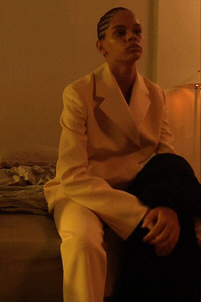 Cream double-breasted blazer with broad peaked lapels and a long clean line. Matching cream trousers with a pressed crease catching the lamplight. Black trouser leg draped across one knee, split look held in the seated pose. Hotel room lamplight after midnight, suitcase still open on the bed.