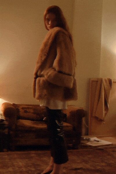Honey-brown fur coat with a deep shawl collar and belted waist. Pale tunic hem peeking below the coat. Black cropped trousers with a lacquered shine at the thigh. Pointed black flats. In a borrowed apartment after midnight, lamps left on in every room.