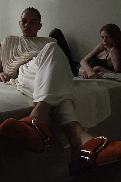 Ivory jersey draped in deep cowl folds across one shoulder. Matching ivory trousers, pooling softly over the ankle. Rust suede slides with oversized gold horsebit hardware. Morning after a hotel party, curtains still closed against the heat.