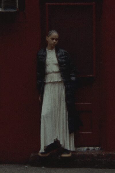Black quilted puffer jacket, cropped high over the waistline. Ivory pleated dress with a dropped ruffle seam and long fluid skirt. Dark shoes with thick pale soles. Outside a red door after rain, holding your coat closed against the cold.