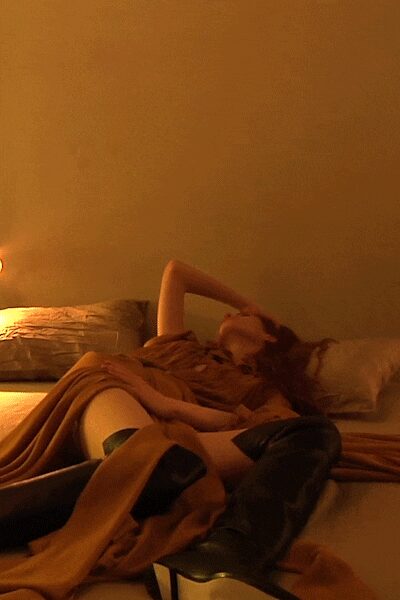 Tobacco jersey dress draped into loose folds across the lap. Black knee-high leather boots with a hard almond toe. Hotel room lamplight, after midnight and not ready to sleep.
