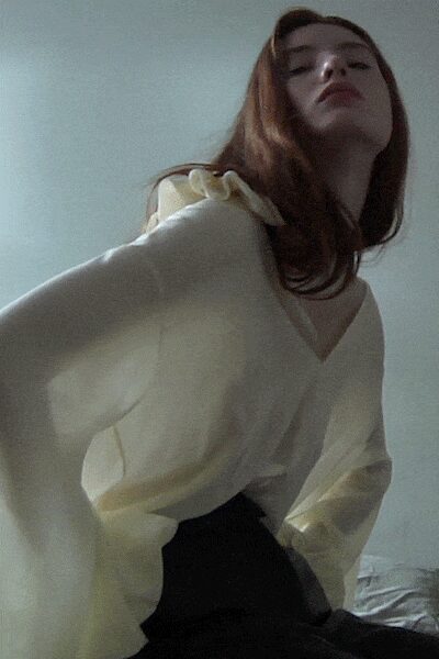 Ivory blouse with a deep V and loose draped sleeves. Soft shoulder ruffle lifting slightly at one side. Black high-waisted skirt, smooth and close through the hip. Afternoon light in a borrowed apartment, before anyone finds the keys.