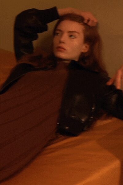 Brown ribbed knit dress falling long and close through the body. Black leather jacket shrugged low, zip teeth catching the warm light. Still on the hotel bed after taking off only half the outfit.