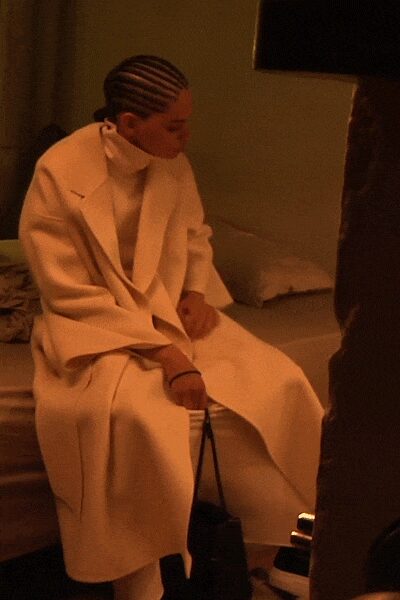 Long ivory wool coat with broad lapels and dropped shoulder seams. Ivory knit turtleneck pulled high against the jaw. Wide ivory trousers breaking softly over the shoe. Small black box bag on thin double handles. Hotel room lamplight, sitting down before taking off one last layer.
