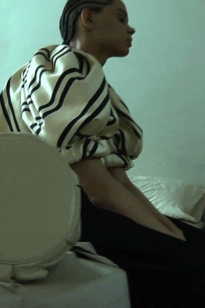 Cream draped top with thick black wavy stripes, sleeves bunched at the elbow. Black slim trousers disappearing into the seat line. Structured ivory shoulder bag with a rounded flap. Sitting in a rented apartment before dawn, still dressed from somewhere louder.
