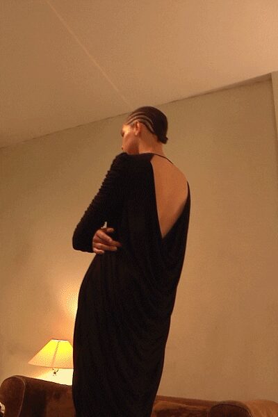 Striped close-fit headscarf wrapping the head like a swim cap. Black jersey column dress with a deep draped back to the waist. Hotel lamp glow, turning away mid-conversation on someone else's sofa.
