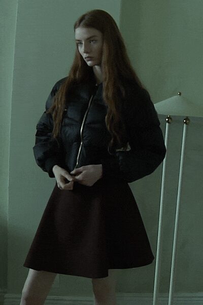 Black cropped puffer jacket with a pale zip running straight off-center. Dark oxblood flared skirt in stiff cloth, hem holding a clean bell. Small ring glinting at one finger. In a rented room before dawn, radiator clicking louder than your thoughts.