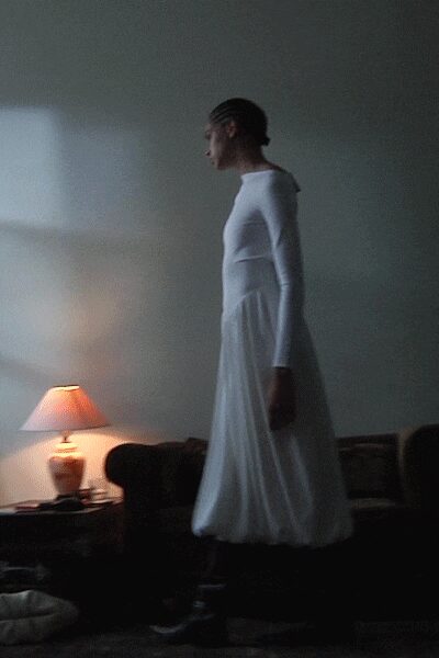 White jersey dress with fitted long sleeves and a softly dropped waist seam. Dark ankle boots under the gathered calf-length hem. Standing on a sofa to reach the window latch after dusk.