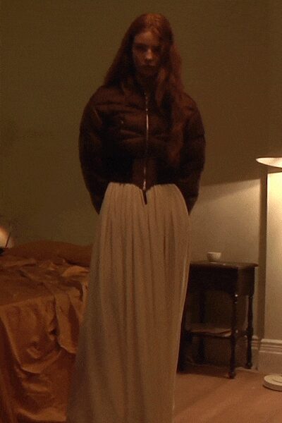 Dark brown padded jacket, cropped hard at the ribs with a silver zip. Chalk-white pleated skirt falling from a gathered waist to the floor. Hotel room lamp glow, still dressed from dinner and not ready to sleep.