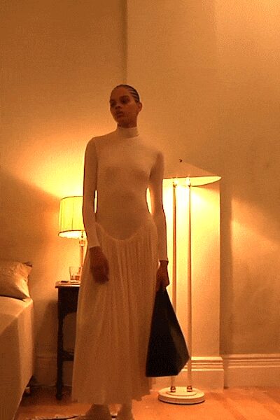 Ivory second-skin bodysuit with a mock neck and long narrow sleeves. Sheer white gathered skirt floating from a dropped waist seam. Large black triangular tote, stiff against the calf. Hotel room lampglow, dressed before dawn while the hallway stays silent.