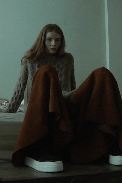 Grey cable-knit sweater with thick braided sleeves and a close mock neck. Rust wool skirt pooled over the knees in heavy folded panels. White platform soles peeking clean beneath the hem. First hour in a rented room after the heat has clicked off.