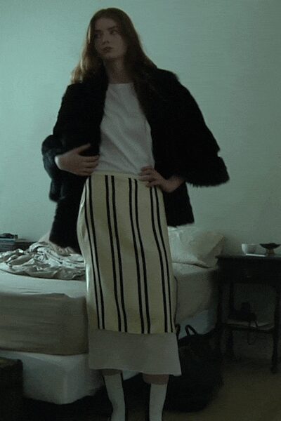 Black fuzzy jacket with a broad, shaggy shoulder line. White crewneck top, plain and softly draped under the jacket. Cream column skirt striped in black, with a deep plain hem. Opaque white tights. Hotel room morning, coffee gone cold on the side table.
