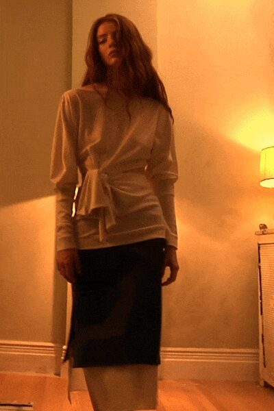Pearl satin top with a twisted side tie and long cuffed sleeves. Black pencil skirt, sheer panel ghosting over a pale underlayer. Pointed black pumps. Lamp-lit hotel room, deciding whether to go back downstairs.