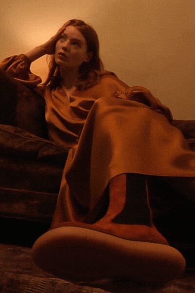 Ochre satin dress with dropped shoulders and a loose fold at the waist. Tall sock boots in rust suede with thick pale rubber soles. Apartment heat turned too high, sunset already gone orange on the walls.