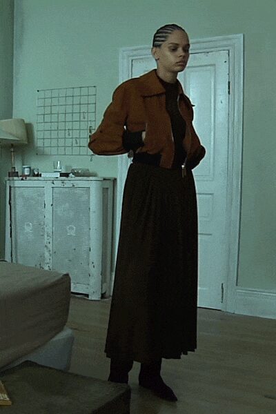 Rust cropped jacket with broad collar and gathered sleeves. Black high-neck underlayer disappearing into the waist. Black pleated midi skirt falling heavy from a cinched waist. Pointed black boots mostly lost under the hem. In a rented room before dawn, radiator ticking louder than your thoughts.