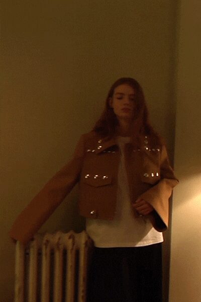 Cropped tan jacket with silver studs tracing the flap pockets. White crewneck layer hanging loose below the jacket hem. Black wide-leg trousers falling in a flat dark column. Hallway radiator ticking after midnight, one lamp left on.