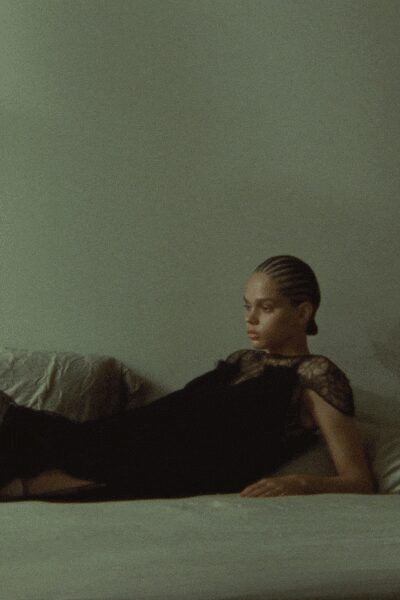 Black column dress with sheer lace yoke and cap sleeves. Short gold chain at the collarbone. Small gold studs. Hotel bed at noon, curtains barely open after a wedding.