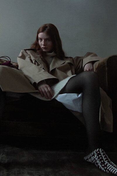 Stone trench slouched open, broad lapels folding into each other. White dress spilling from under it in a stiff triangular panel. Charcoal sheer tights. Black-and-white pumps in a warped checker pattern. Sinking into a hotel lobby sofa after the rain started again.