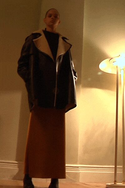 Oversized black leather shearling jacket with broad cream lapels and a skewed zip. Black high-neck layer, close and plain under the heavy outer shell. Rust column skirt falling straight to the ankles without a break. Black pointed boots, mostly swallowed by the hem. Standing in a rented flat lit only by the floor lamp.
