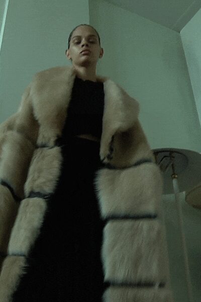 Oversized blush fur coat banded with narrow black horizontal strips. Black cropped top, cut close and plain under the volume. Long black skirt falling straight from beneath the cropped hem. In a cold stairwell after midnight, keys still somewhere in the bag.