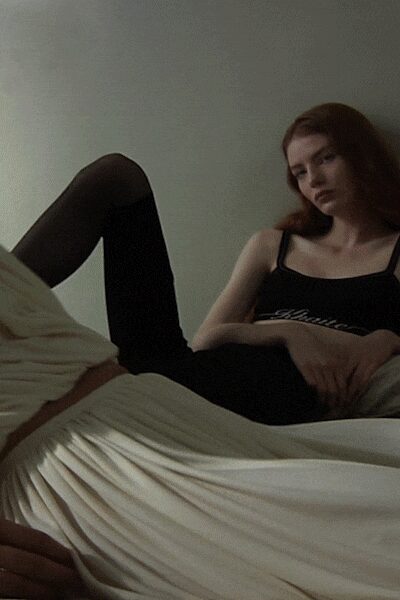 Black jersey slip dress with narrow straps and a low scooped neckline. Sheer black tights, one knee lifted into the light. Ivory pleated skirt spread across the bed, held with a tan belt. Afternoon in a rented room, the sheets still creased from a nap.
