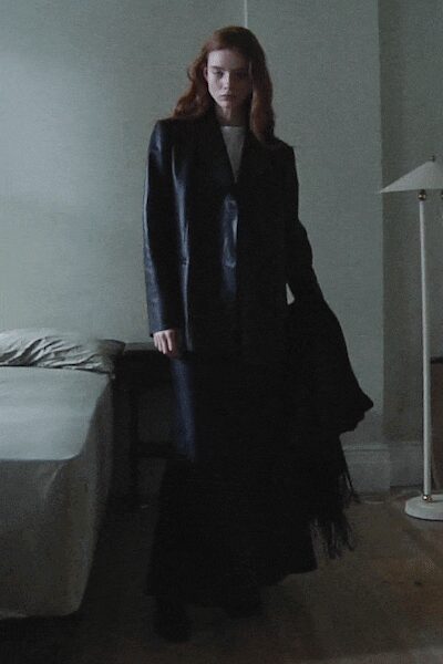 Long black leather blazer with a narrow notch lapel. White crewneck underlayer, flat and plain at the collar. Floor-length black skirt, dissolving into a soft tulle haze. Black pointed boots barely breaking the hem. In a rented room after checkout, lamp still on and shoes back on.