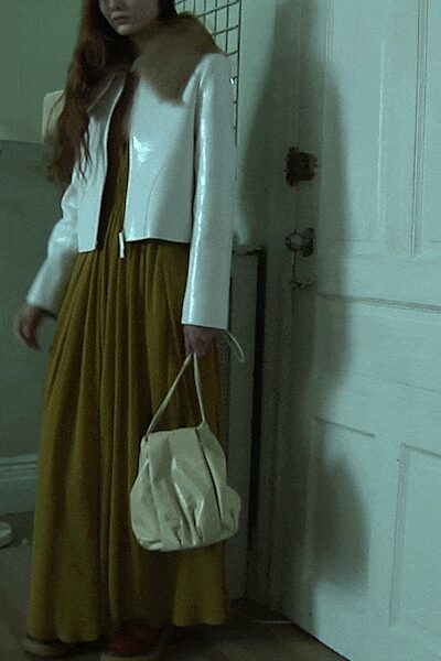 White patent jacket with a soft brown fur collar and dangling ties. Ochre jersey dress falling in fine gathers from under the bust. Cream drawstring bucket bag, slouched and lightly puckered. Rust flats just visible below the hem. By the apartment door, deciding whether the rain is worth going back out.