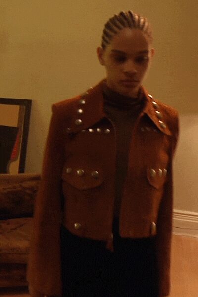 Cropped rust suede jacket, silver dome studs marching over the yoke and pockets. Dark close-fit turtleneck underlayer. Black trousers falling straight from the hip. In a wood-paneled living room after everyone else has gone upstairs.
